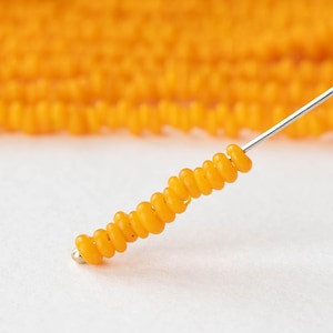 May include: Close-up of a strand of bright orange beads strung on a silver needle. The beads are small, round, and evenly spaced. A blurred background shows more strands of the same beads. The beads are likely for jewelry making or crafting.