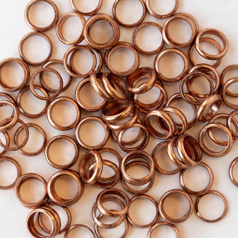 Pu&ograve; includere: Primo piano di numerosi piccoli anelli metallici color rame. Ogni anello &egrave; circolare con un foro centrale. Gli anelli sono sparsi su una superficie bianca e hanno una finitura metallica lucida. L'immagine mostra una collezione di questi anelli.