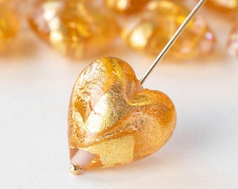 May include: A heart-shaped glass bead with gold leaf accents. The bead is a light amber color and has a translucent finish.