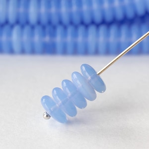 May include: A close-up of light blue, oval-shaped beads strung on a silver needle. The beads are translucent and have a smooth, glossy finish. The background features a blurred array of similar beads, suggesting a larger collection.