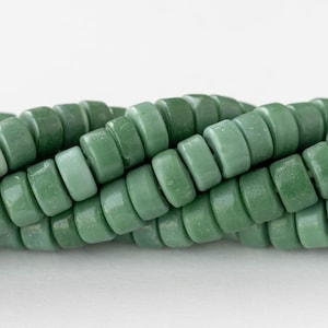 May include: Close-up of a strand of green, disc-shaped beads. The beads have a matte finish and vary slightly in shade, ranging from light to dark green. The beads are strung together, creating a textured, organic look.
