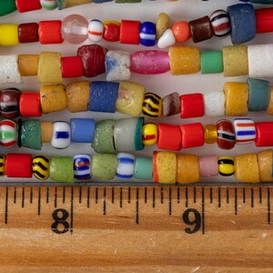 May include: Close-up of several strands of colorful glass beads. The beads are various shapes and sizes, including round, square, and cylindrical. Colors include red, yellow, blue, green, and white. A wooden ruler with inch markings is at the bottom.