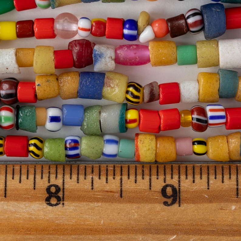 May include: Close-up of several strands of colorful glass beads. The beads are various shapes and sizes, including round, square, and cylindrical. Colors include red, yellow, blue, green, and white. A wooden ruler with inch markings is at the bottom.