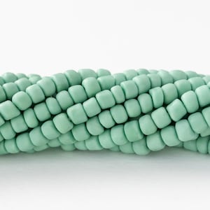 Könnte beinhalten: Nahaufnahme eines Strangs hellgrüner, quadratischer Perlen. Die Perlen sind in einer gebogenen Linie angeordnet, was auf eine Halsketten- oder Armbandkomponente hindeutet. Die Perlen haben eine matte Oberfläche und sind gleichmäßig gefärbt.