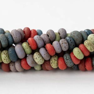 Op de afbeelding: Close-up van een streng kleurrijke, getextureerde kralen. De kralen zijn in verschillende kleuren, waaronder rood, groen, blauw en paars. De kralen zijn ovaal van vorm en hebben een gespikkelde uitstraling. De achtergrond is wit.