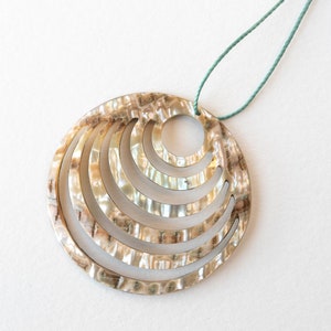 May include: A round, iridescent, mother-of-pearl pendant with a concentric circle design. The pendant is attached to a green cord.