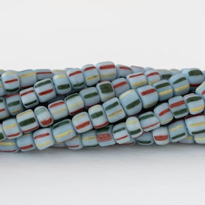 Puede incluir: Primer plano de un collar de cuentas rectangulares de color azul claro. Cada cuenta tiene un color base azul claro con rayas rojas, amarillas y verdes. Las cuentas están ensartadas, creando una superficie texturizada.