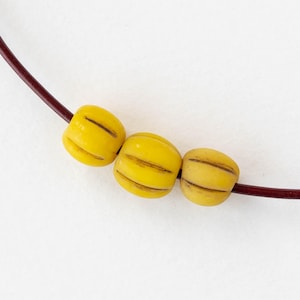 May include: A close-up of a necklace featuring three yellow, barrel-shaped beads with brown stripes. The beads are strung on a dark red cord, set against a white background. The necklace is a simple, yet elegant piece of jewelry.