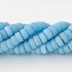 May include: Close-up of strands of light blue, disc-shaped beads. The beads are strung together, creating a wavy pattern. The beads have a matte finish and are evenly sized. The background is white.