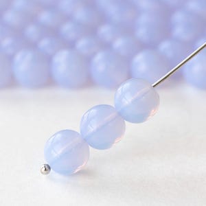 Puede incluir: Primer plano de tres cuentas redondas de color azul pálido ensartadas en una aguja plateada. Las cuentas tienen una cualidad translúcida, con sutiles variaciones de color. El fondo presenta más cuentas desenfocadas, lo que sugiere una colección más grande.