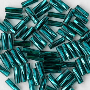 May include: A collection of teal-colored, cylindrical glass beads. The beads have a metallic sheen and are scattered across a white surface. The beads are approximately 0.25 inches in length, suitable for jewelry making and crafts.