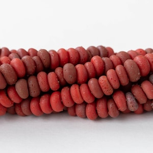 May include: Close-up of strands of red and maroon beads. The oval-shaped beads have a slightly textured surface. The beads are strung together, creating a layered effect. The background is plain white.