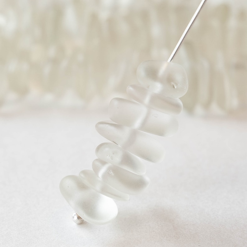 Pu&ograve; includere: Una fila di perline di vetro bianco smerigliato, infilate su un filo d'argento. Le perline sono impilate in dimensioni graduate, con la perlina pi&ugrave; grande in basso e la perlina pi&ugrave; piccola in alto.