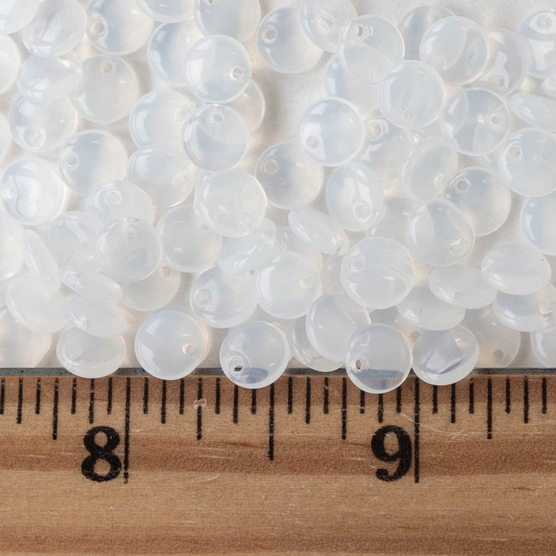May include: A close-up of a pile of white, translucent, round, flat beads with a hole in the center. The beads are arranged in a random pattern and are about 1 inch in diameter. A wooden ruler with inch markings is visible at the bottom of the image.