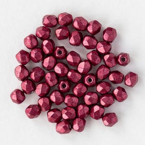 May include: A selection of faceted, metallic burgundy beads. The beads have a geometric shape with multiple flat surfaces. The beads are scattered on a white surface, highlighting their colour and texture. These beads are suitable for jewellery making and crafting.
