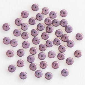 May include: A collection of metallic purple disc-shaped beads with a central hole. The beads have a slightly textured surface and a bronze-colored sheen, scattered on a white background. These beads are suitable for jewelry making and crafting projects.