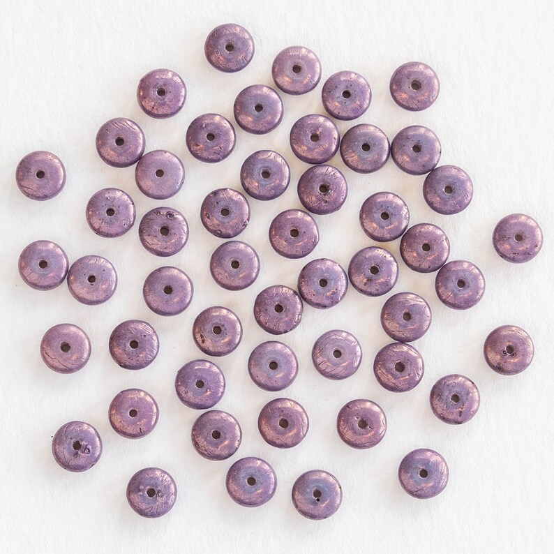 May include: A collection of metallic purple disc-shaped beads with a central hole. The beads have a slightly textured surface and a bronze-colored sheen, scattered on a white background. These beads are suitable for jewelry making and crafting projects.