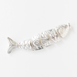 May include: A silver-colored fish-shaped object with segmented body. The fish has detailed scales and fins, and is lying on a white surface. The fish is a decorative item.