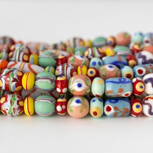 Può includere: Primo piano di una collezione di perline di vetro colorate in varie forme e dimensioni. Le perline presentano un mix di colori tra cui rosso, giallo, verde, blu e arancione, con motivi e disegni intricati. Le perline sono disposte vicine.