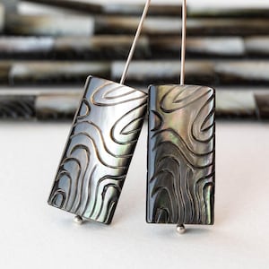 May include: Two black and iridescent rectangular earrings with a carved wave pattern. The earrings are hanging from silver posts.