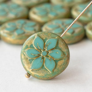 May include: A close-up of a teal blue glass bead with a gold flower design. The bead is round and has a smooth, glossy finish.