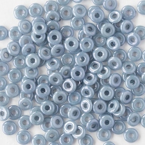 May include: A pile of light blue, ring-shaped glass beads. The small beads have a hole in the middle and seem to be made of a translucent material. The beads are scattered on a white surface.