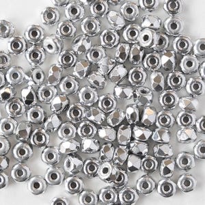 May include: A collection of small, shiny silver beads. The beads have a variety of shapes, including round and faceted, with a hole through the center for stringing. The beads are scattered across a white surface.