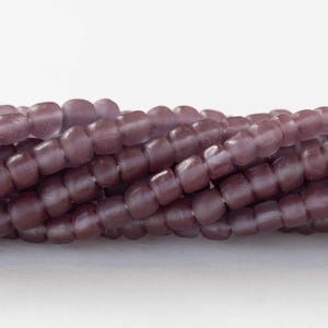 May include: A strand of small, opaque, purple glass beads. The beads are round and have a slightly irregular shape. They are arranged in a loose pile.