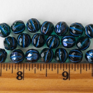 May include: A row of dark teal blue glass beads with a dark blue stripe around the center of each bead. The beads are arranged in a line on a white surface with a wooden ruler below them.