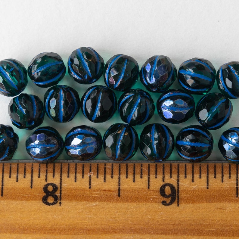 May include: A row of dark teal blue glass beads with a dark blue stripe around the center of each bead. The beads are arranged in a line on a white surface with a wooden ruler below them.