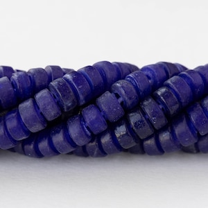 May include: Close-up of a strand of dark blue, disc-shaped beads. The beads are strung together, creating a textured, rope-like appearance. The beads have a slightly irregular shape and a matte finish, with visible imperfections.