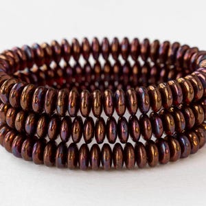 May include: Close-up of three stacked bracelets made of small, iridescent, brown beads. The beads have a metallic sheen with hints of purple and gold. The bracelets are arranged in a spiral pattern.