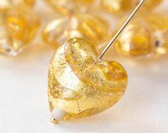 May include: Close-up of heart-shaped beads with gold leaf inclusions. A gold needle pierces one bead, showcasing the translucent quality. The beads are a warm gold color, ideal for jewelry making.