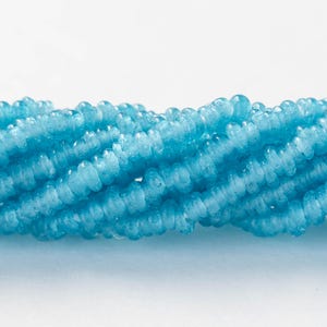 Puede incluir: Primer plano de un hilo de cuentas de piedras preciosas azul claro. Las cuentas son pequeñas, redondas y tienen una superficie ligeramente texturizada. Las cuentas están ensartadas, creando una forma fluida y orgánica. El fondo es blanco liso, resaltando el color y la textura de las cuentas.
