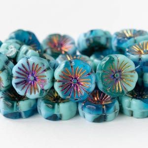 May include: Close-up of a collection of turquoise flower-shaped beads. Each bead has a central design with gold and purple accents. The beads are clustered together, showcasing their intricate details and color variations.