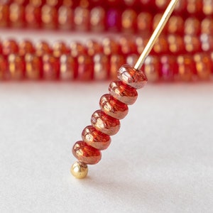 May include: Close-up of a gold wire with a stack of small, round, iridescent red beads. The beads have a metallic sheen and are strung on a gold wire.