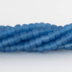 May include: Close-up of strands of blue glass beads. The beads are cylindrical with rounded edges and a matte finish. The beads are strung together, creating a textured, organic pattern. The color is a muted, dusty blue.
