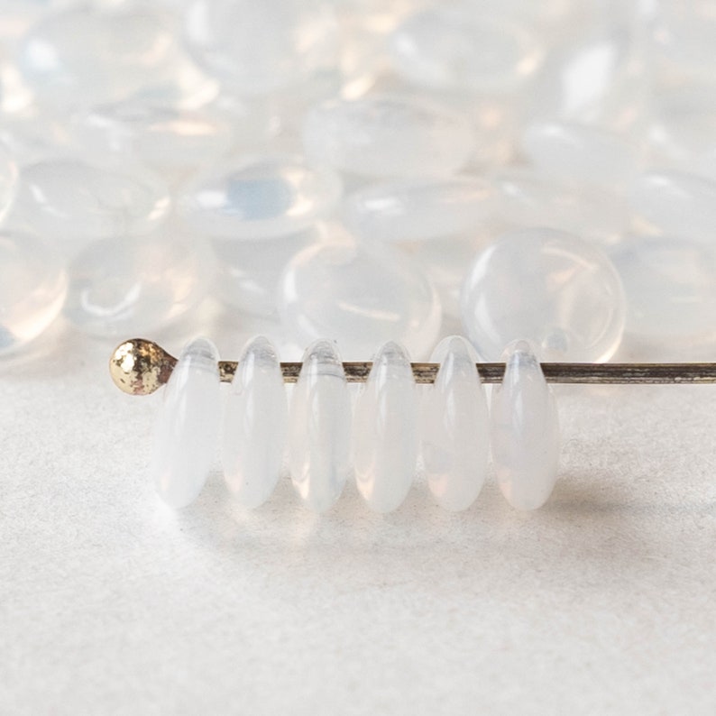 May include: A strand of white oval-shaped glass beads strung on a silver wire. The beads are translucent and have a slight iridescent sheen.
