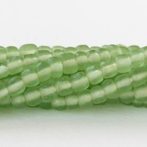 Pode incluir: Um fio de contas de vidro verde claro com acabamento fosco. As contas são quadradas e têm uma textura ligeiramente irregular.