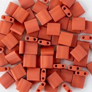 Può includere: Una collezione di perline quadrate e rettangolari rosso-arancio. Le perline sono realizzate in un materiale opaco e hanno un colore uniforme. Alcune perline hanno due fori per l'infilatura. Le perline sono sparse su una superficie bianca.
