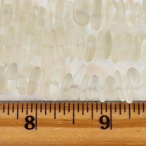 Pu&ograve; includere: Un filo di perline di vetro bianco traslucido, alcune ovali e altre pi&ugrave; allungate, con un righello che mostra che le perline misurano circa 1,2 cm di lunghezza.