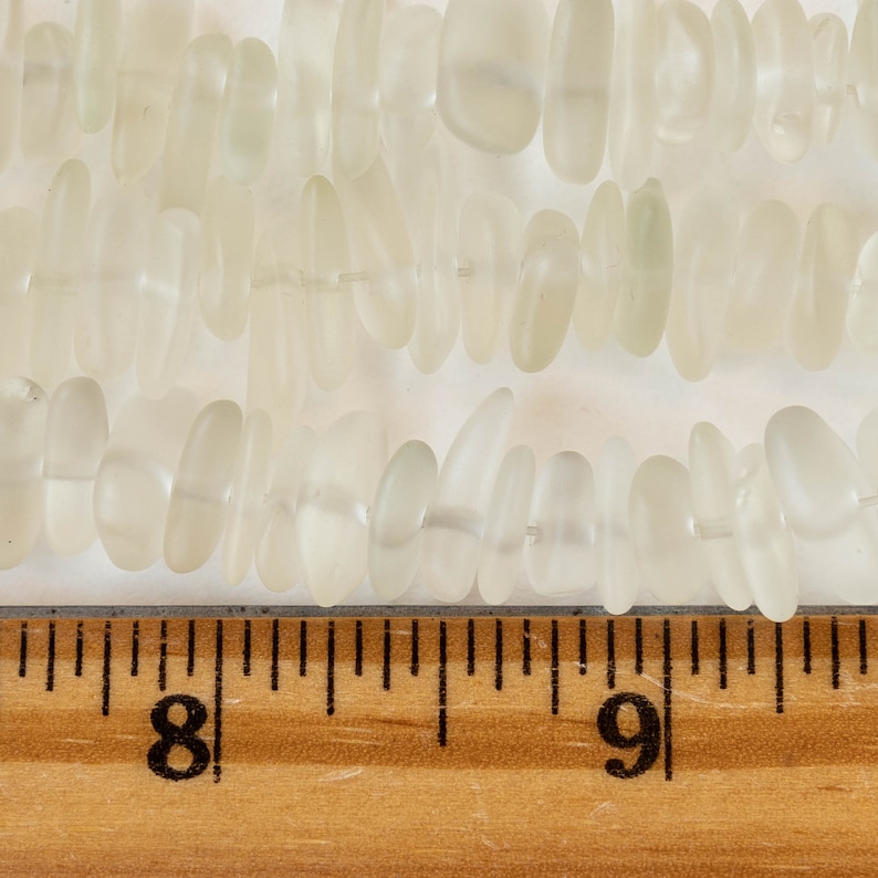 Pu&ograve; includere: Un filo di perline di vetro bianco traslucido, alcune ovali e altre pi&ugrave; allungate, con un righello che mostra che le perline misurano circa 1,2 cm di lunghezza.