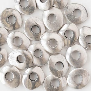 May include: A collection of silver-colored, irregular-shaped metal beads. Each bead has a central hole, and the surfaces have a slightly hammered texture. The beads are scattered, creating a visually appealing arrangement for jewelry making or crafting.