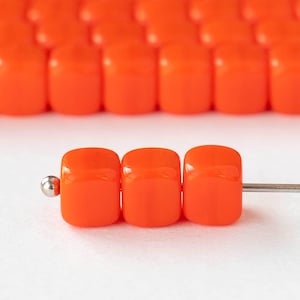 May include: Close-up of bright orange, square-shaped beads strung on a silver needle. The beads have a glossy finish and are arranged in rows, with a shallow depth of field. The beads are likely for jewelry making or crafts.