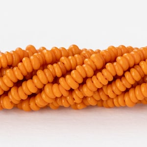Puede incluir: Primer plano de hebras de cuentas de color naranja. Las cuentas son pequeñas, redondas y tienen forma de espiral. Las cuentas están ensartadas, creando un patrón texturizado y visualmente atractivo. El fondo es blanco liso, lo que resalta las cuentas.
