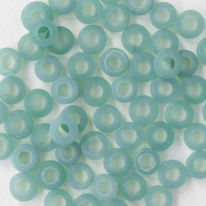 Könnte beinhalten: Eine Sammlung hellblauer, ringförmiger Perlen. Die Perlen sind durchscheinend und haben ein mattiertes Aussehen. Sie sind auf einer weißen Oberfläche verstreut und bilden ein Muster aus Kreisen. Diese Perlen eignen sich für die Schmuckherstellung und Bastelprojekte.