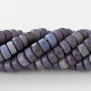 May include: Close-up of a strand of purple and gray disc-shaped beads. The beads vary in shades of purple and gray, creating a gradient effect. The beads are strung together, suggesting they are for jewelry making or crafting.
