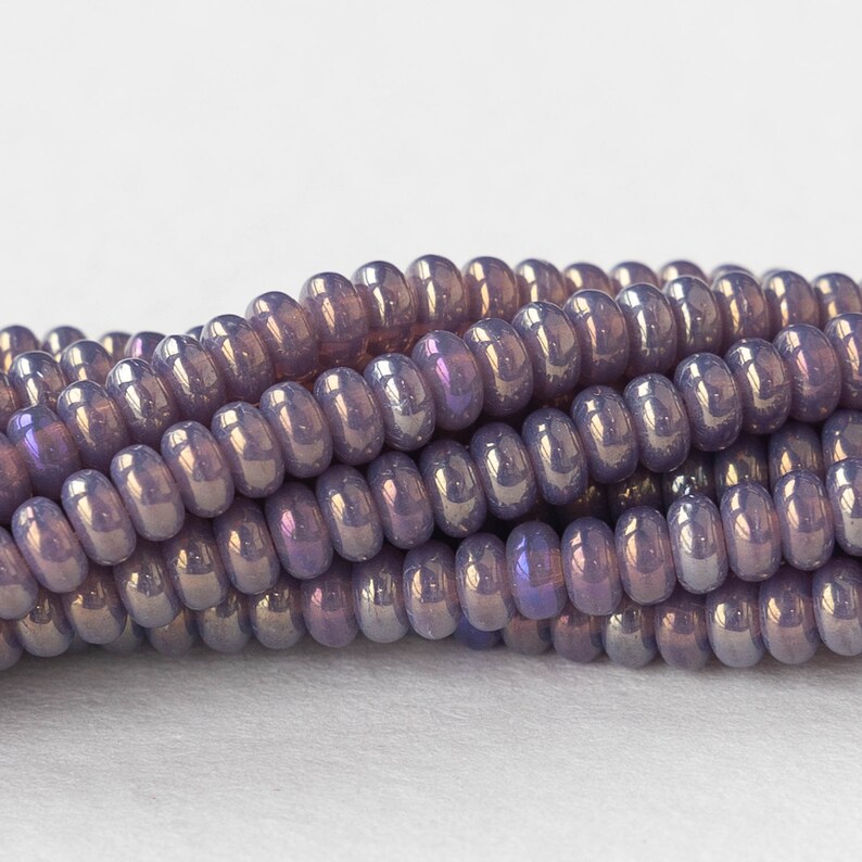 Pu&ograve; includere: Un filo di perline di vetro viola iridescente. Le perline sono piccole e rotonde, con una forma leggermente appiattita. Hanno una finitura lucida e un leggero luccichio.