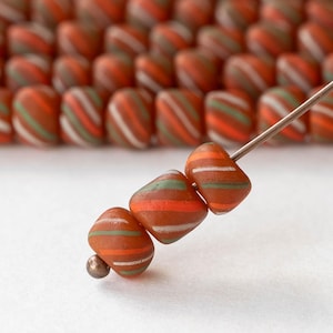 May include: Close-up of three orange, green, and white striped beads on a metal rod. The beads are a reddish-brown colour and have a geometric shape. A row of similar beads is in the background, out of focus. The beads are likely for jewellery making.