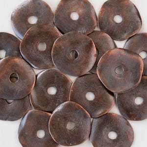 May include: A collection of irregularly shaped, bronze-colored metal discs with a central hole. The discs have a slightly textured surface and vary in size and shape, creating a rustic aesthetic. The arrangement is scattered, showing the details of each piece.